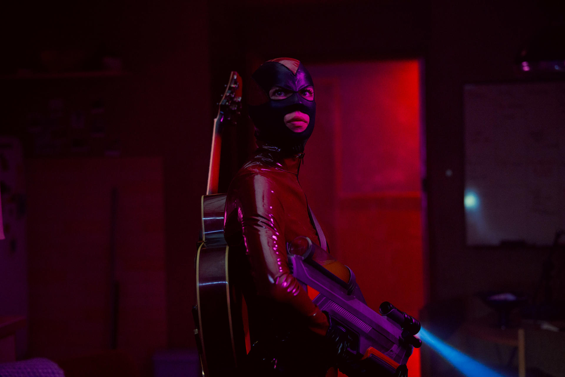 Eine Person im roten Lackanzug und Maske hält eine Waffe und eine Gitarre.