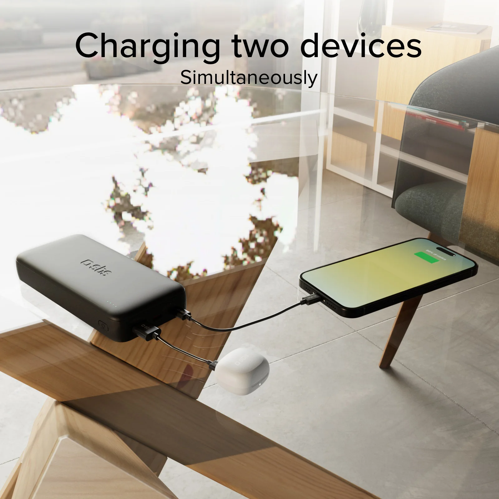 Eine schwarze Powerbank auf einem Glastisch lädt gleichzeitig ein Smartphone und kleine weiße Ohrhörer.