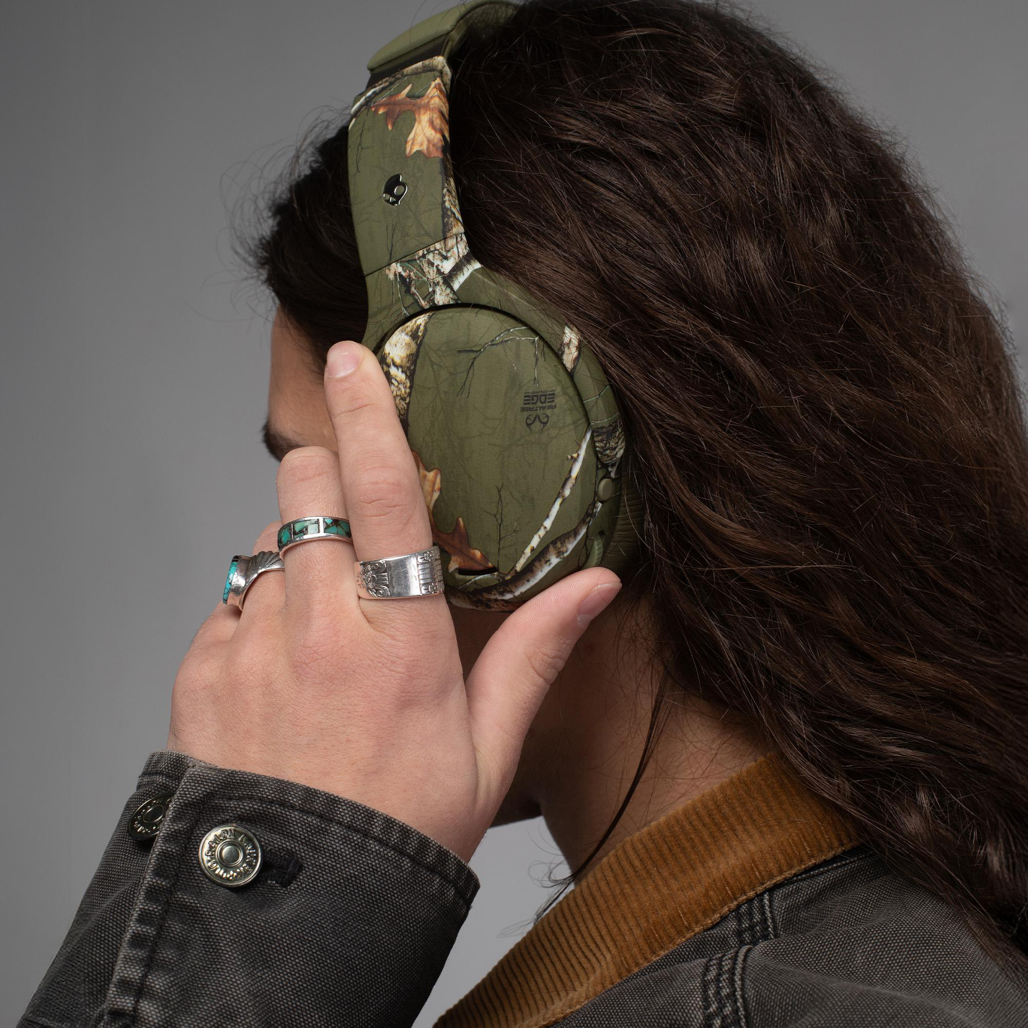 Person mit langem braunen Haar trägt Camouflage-Kopfhörer und Ringe an der Hand.