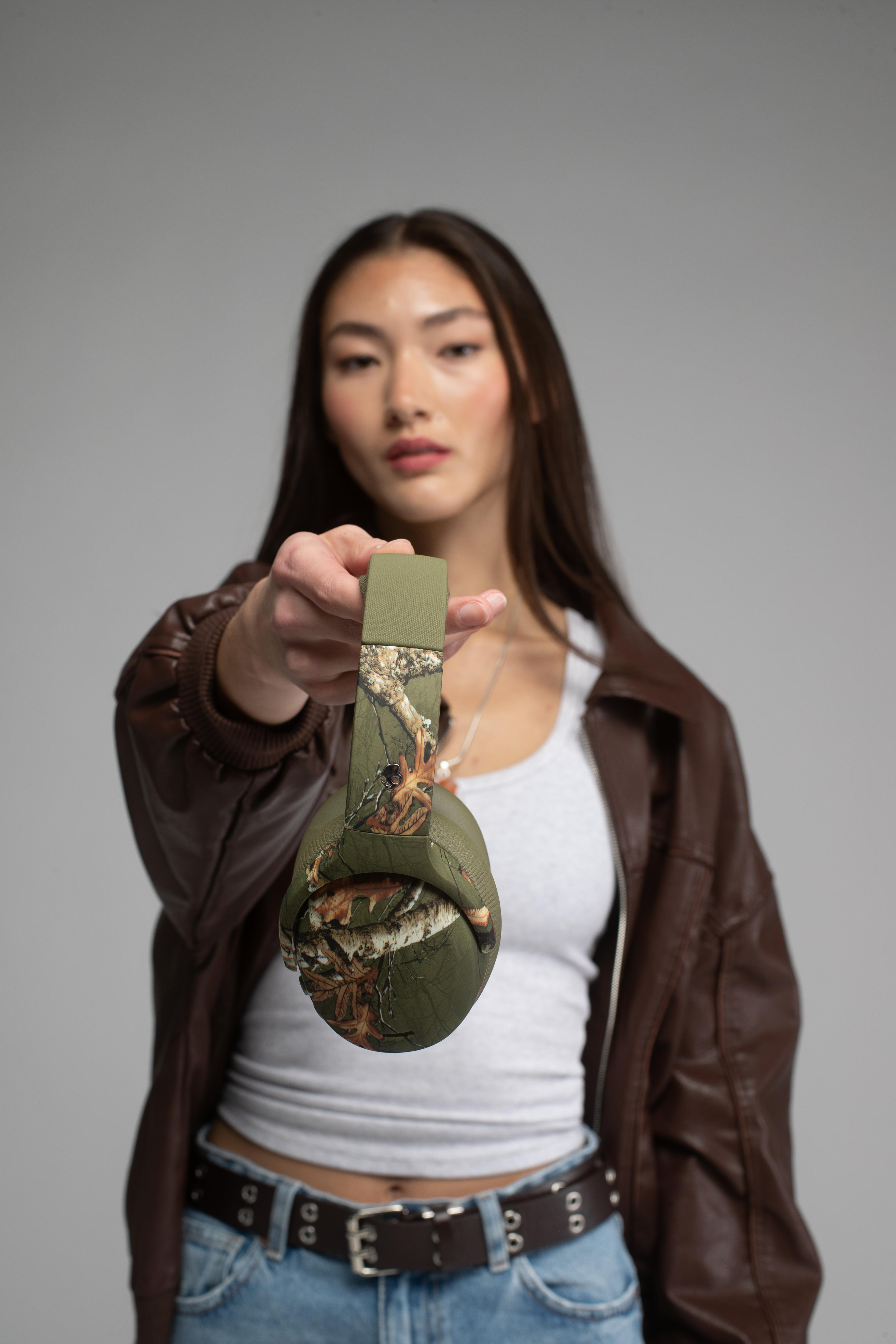 Eine Frau hält grüne Camouflage-Kopfhörer in die Kamera, sie trägt eine braune Lederjacke und ein weißes Tanktop.