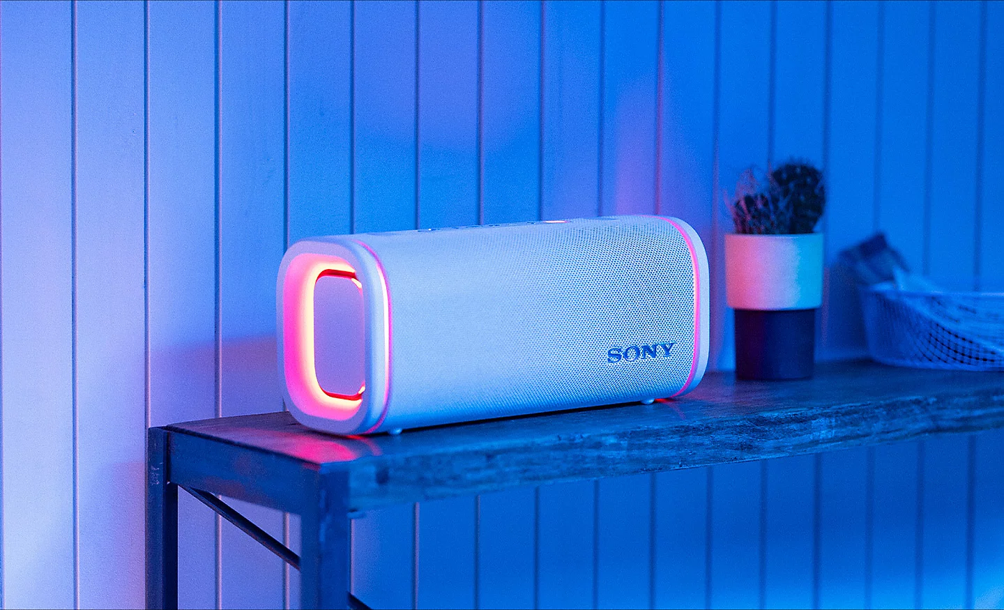 Kırmızı yanan kenarlara sahip beyaz bir Sony Bluetooth hoparlör mavi ahşap duvar önünde duruyor.