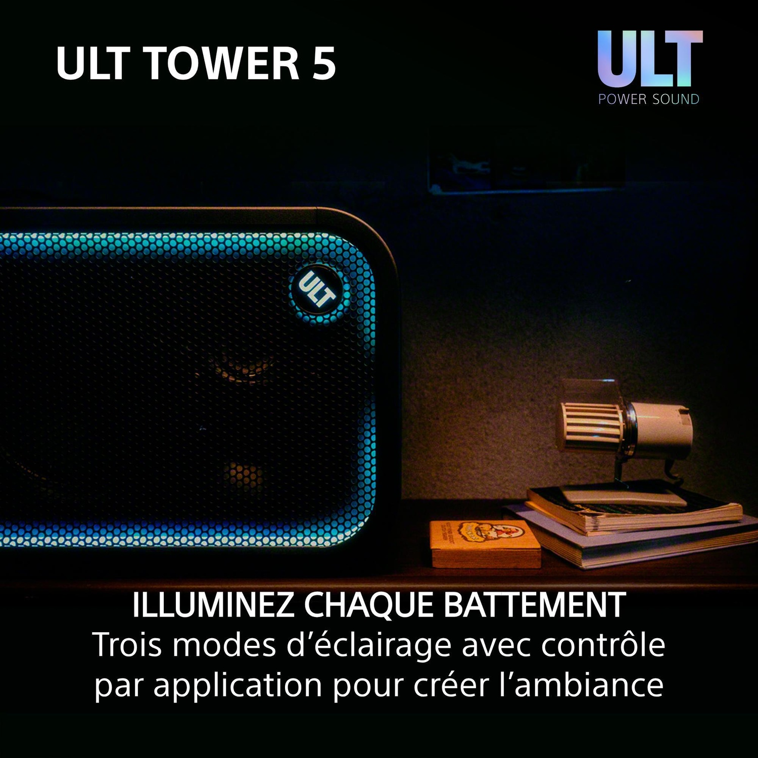 Une enceinte ULT Tower 5 avec éclairage bleu est posée sur un bureau en bois près de livres et d'une petite lampe.