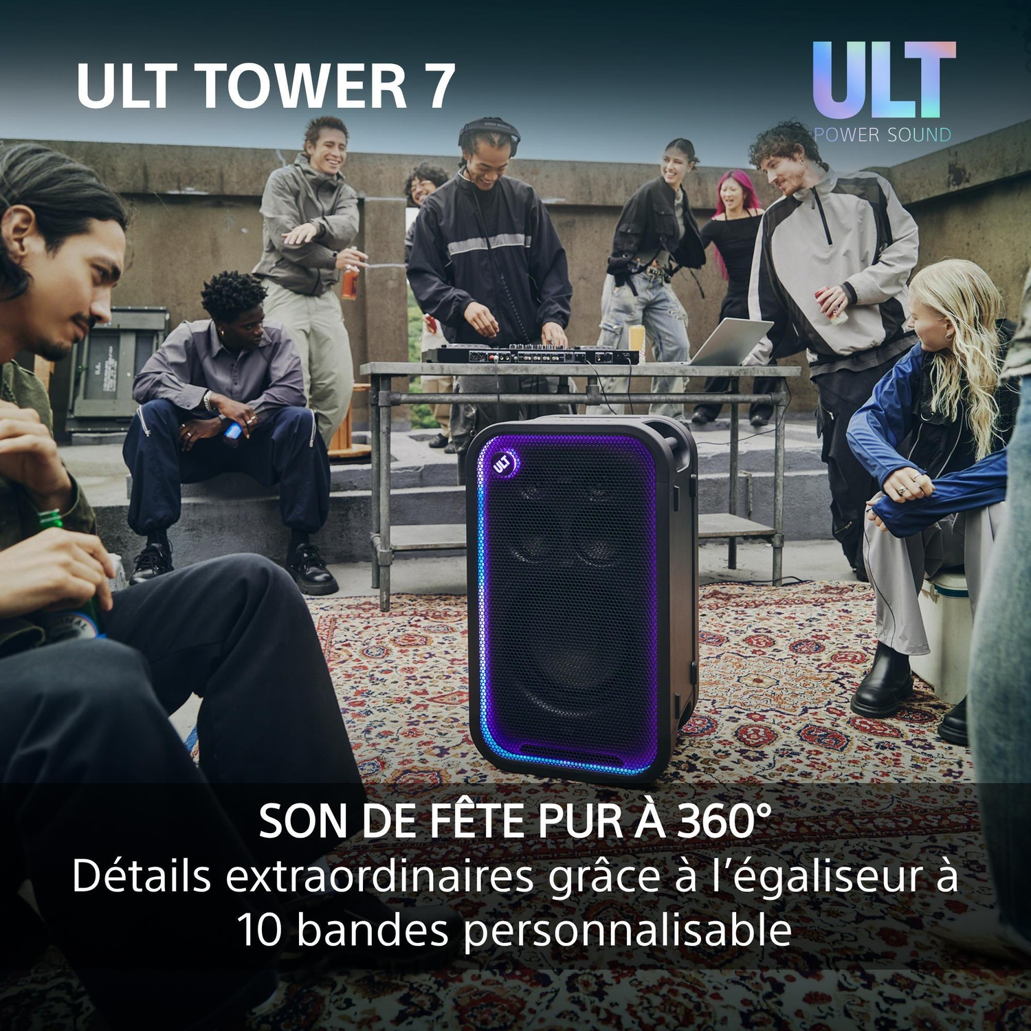 Une grande enceinte portable à éclairage bleu posée sur un tapis, entourée de jeunes sur un toit-terrasse.
