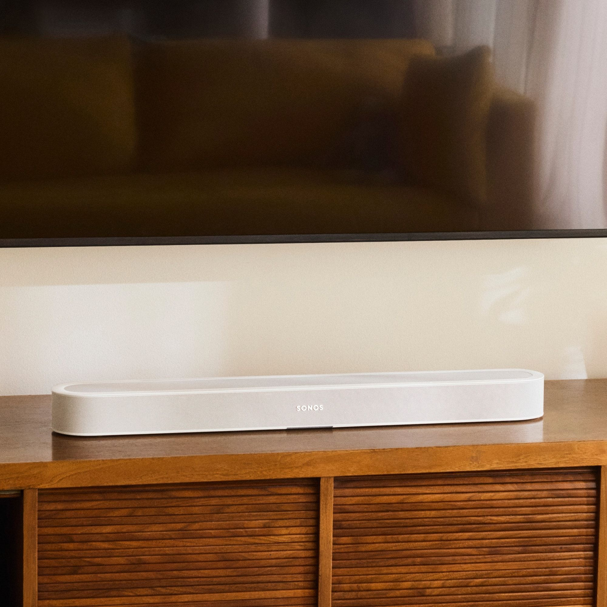 Une barre de son Sonos blanche repose sur un meuble en bois sous un téléviseur.