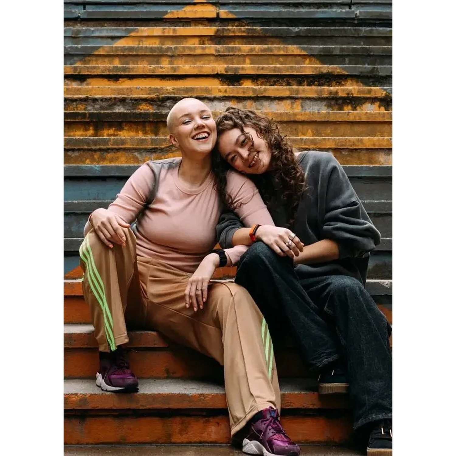 Two smiling young women sitting together on orange stairs, one with a shaved head and the other with curly hair.