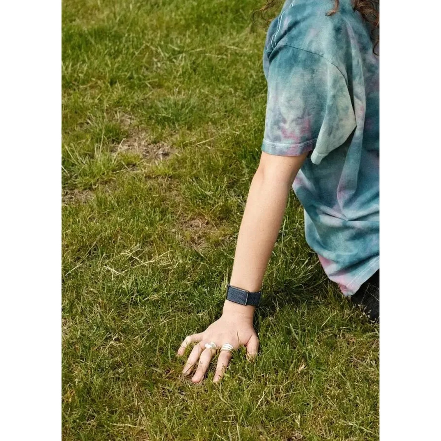 A person in a tie-dye shirt sits on green grass, wearing rings and a dark watch.