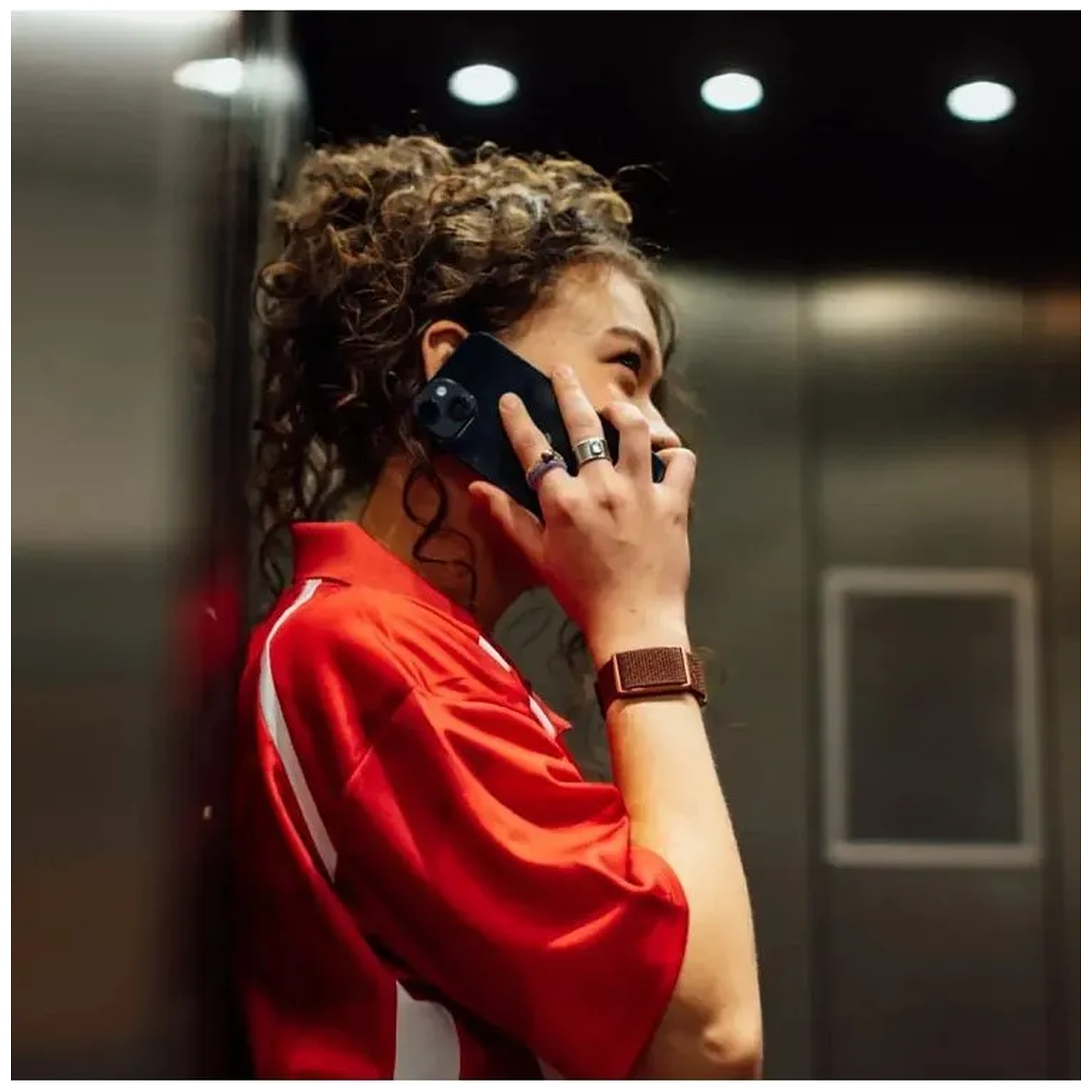 A woman with curly hair wearing a red top is talking on the phone in an elevator.