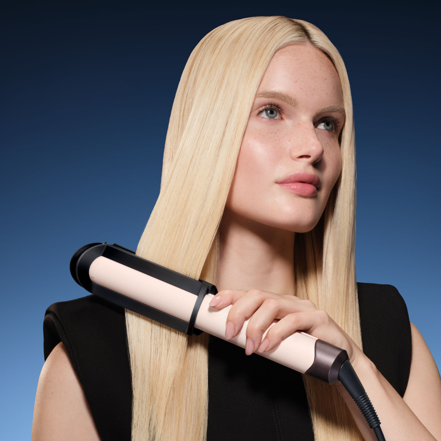 Een blonde vrouw die haar lang, stijl haar stylet met een stijltang tegen een blauwe achtergrond.