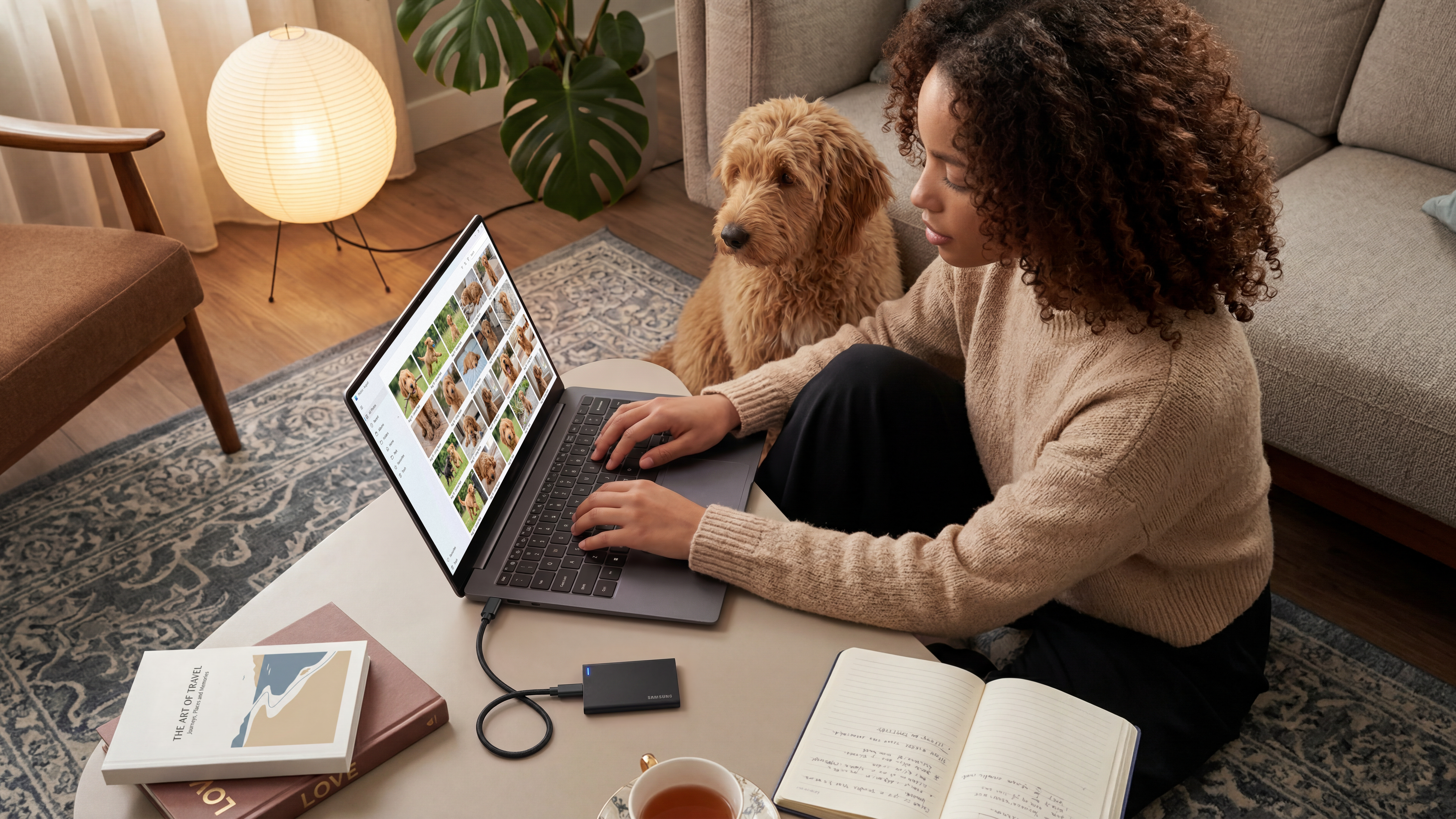 Eine Frau sitzt am Tisch mit einem Laptop und einem Hund, daneben Bücher und Tee.
