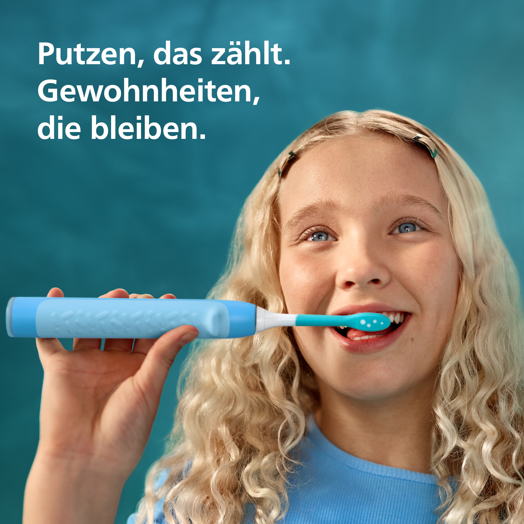 Una ragazza bionda con un top blu si lava i denti con uno spazzolino elettrico blu su sfondo blu.