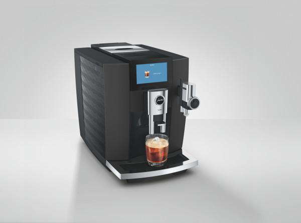 Moderne schwarze Kaffeemaschine mit Display und einer Tasse Kaffee auf einem Tisch vor grauem Hintergrund.
