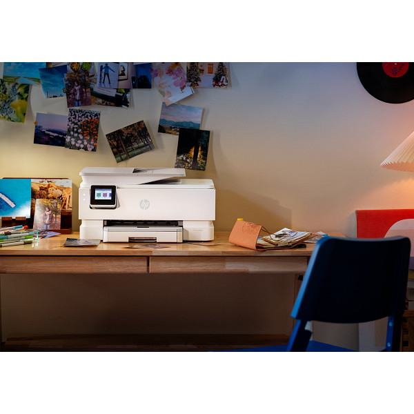 Ein weißer HP Drucker steht auf einem Holztisch, Fotos an der Wand und ein blauer Stuhl im Vordergrund.