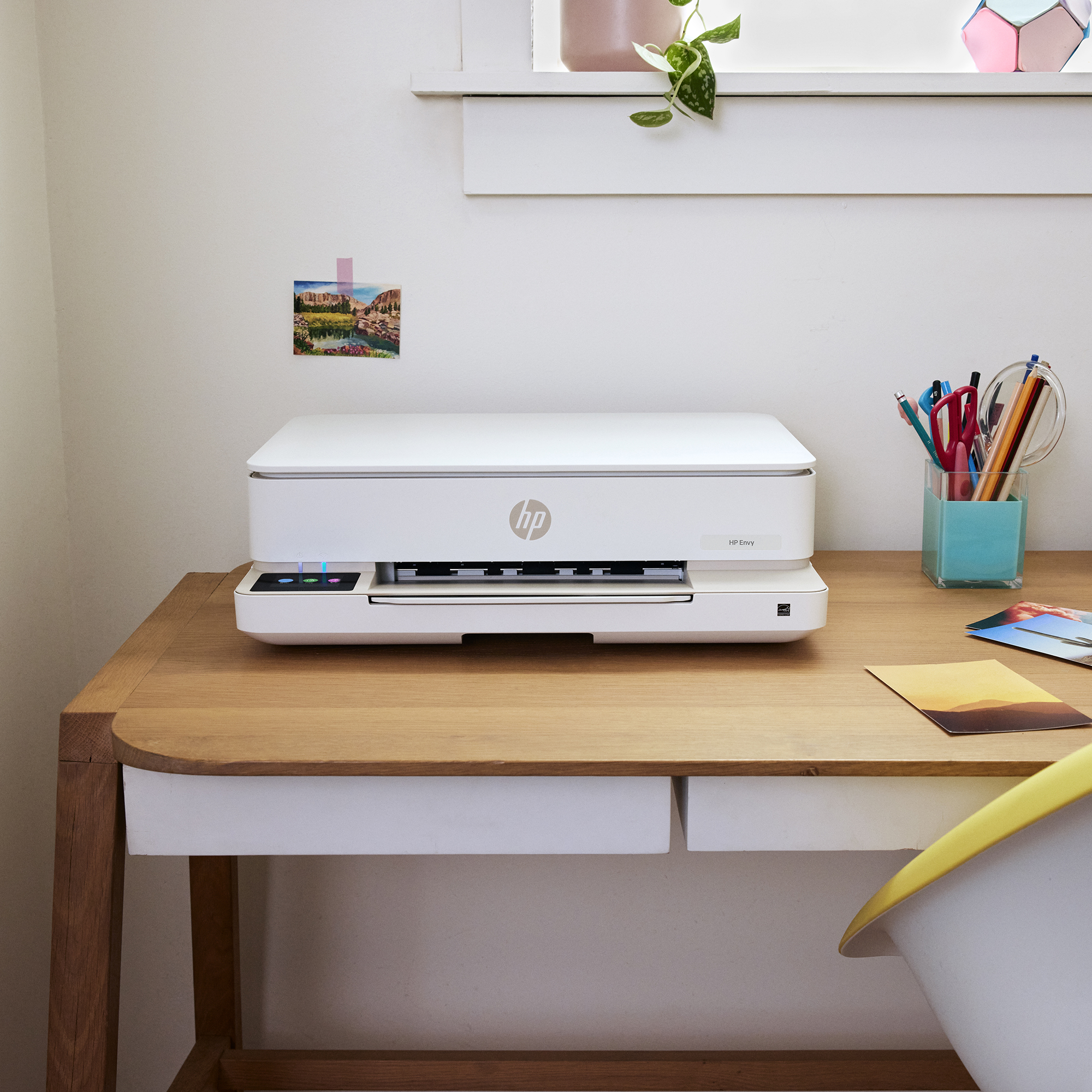 Ein weißer HP Envy Drucker auf einem Holztisch mit Stiften und Fotos daneben.