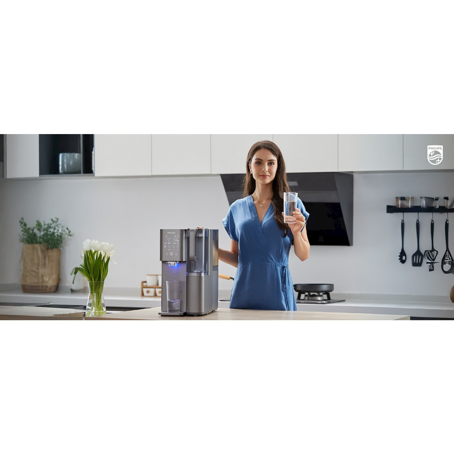 Una donna in abito blu in una cucina moderna tiene un bicchiere d'acqua vicino a un distributore d'acqua Philips.