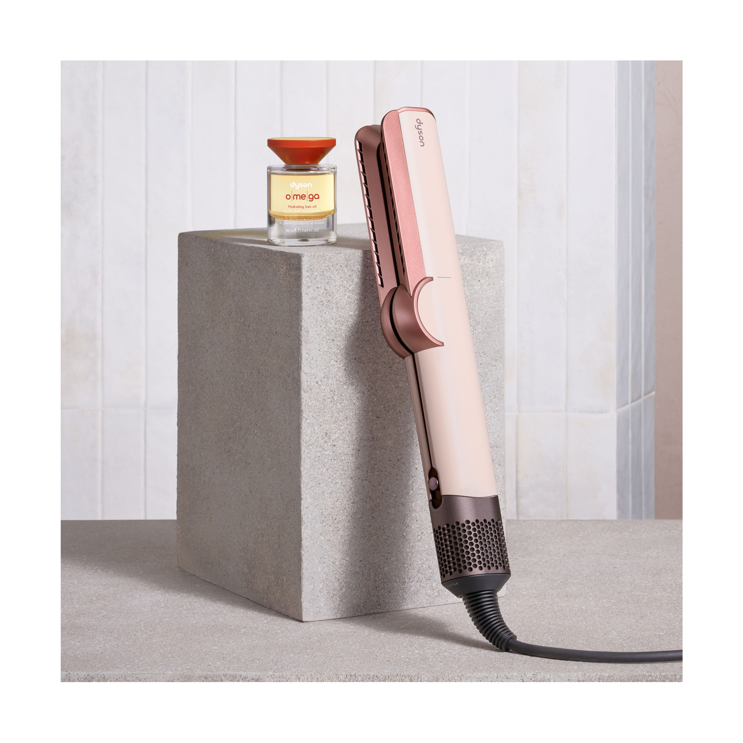 Una piastra per capelli Dyson color oro rosa è in piedi su un piedistallo grigio accanto a una bottiglietta d'olio.