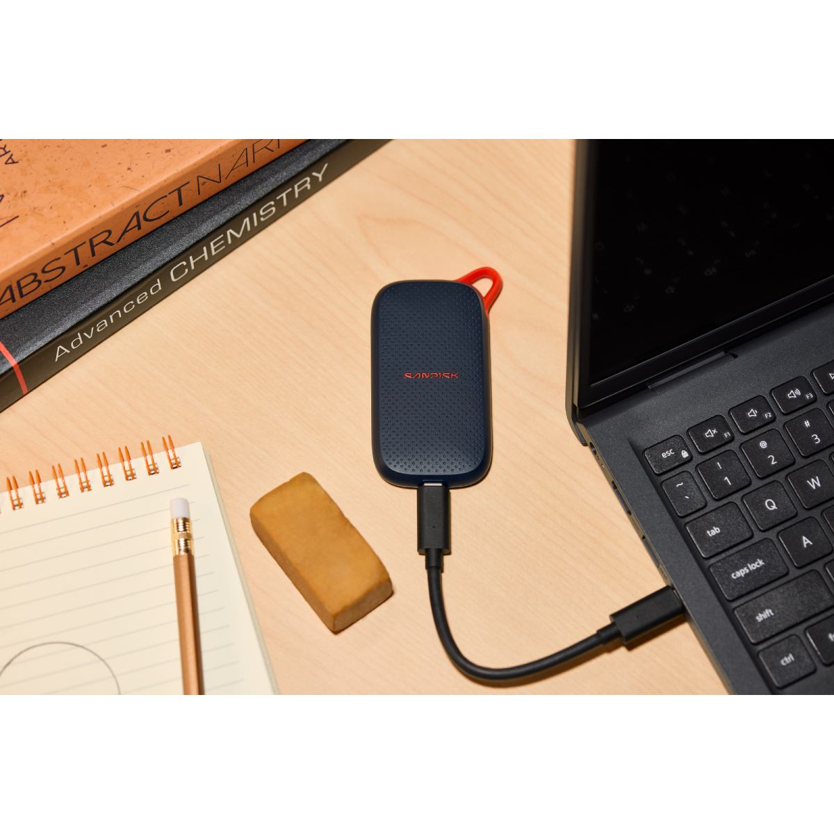 SSD SanDisk azul conectada a una laptop en un escritorio de madera, junto a un cuaderno y libros.