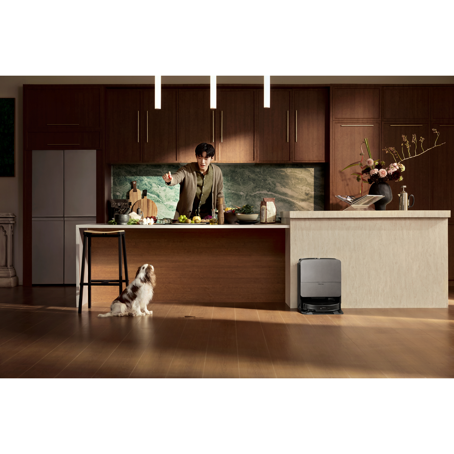 Un homme cuisine dans une cuisine moderne pendant qu'un chien regarde en haut. Un robot aspirateur est sur le sol.