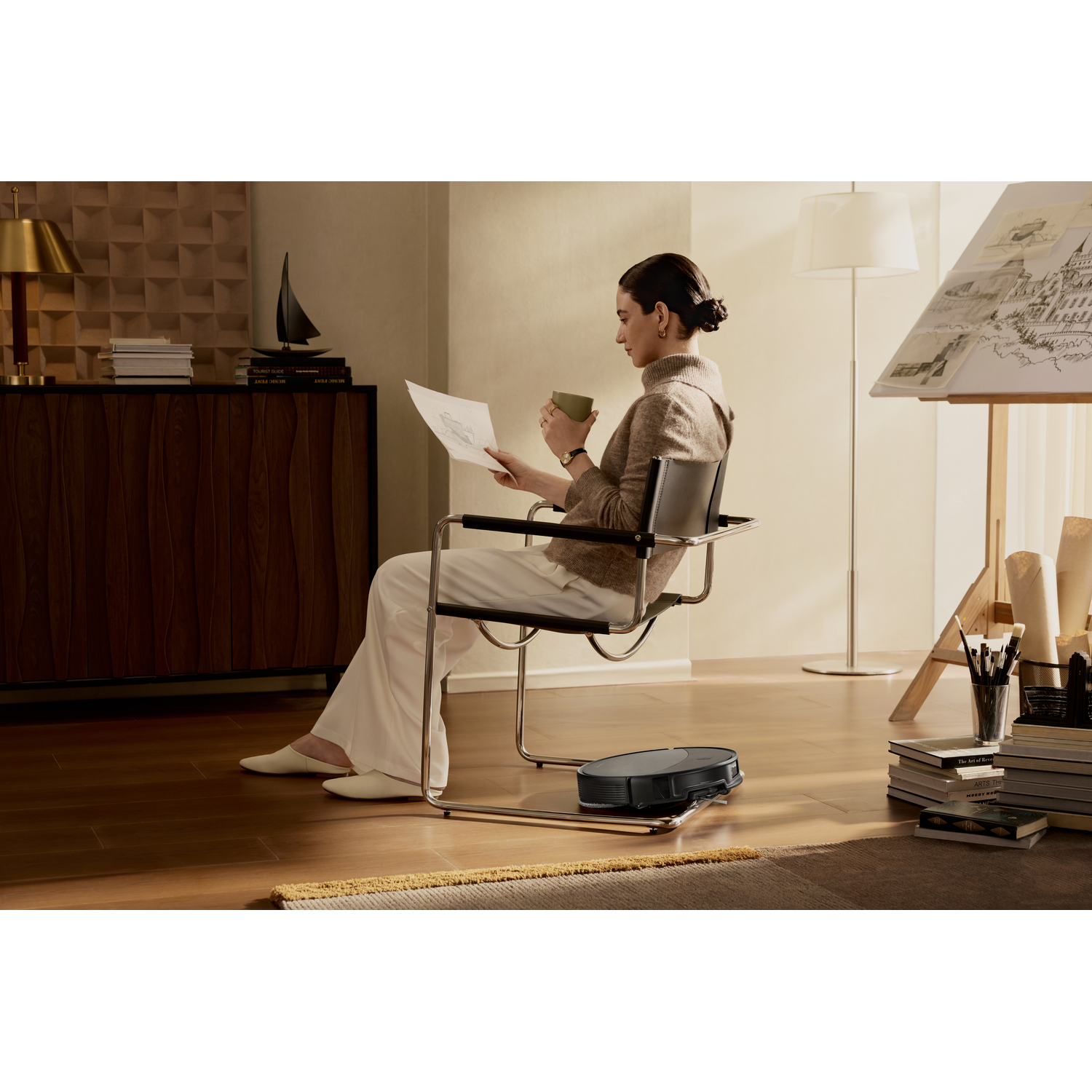Une femme assise lit et boit, tandis qu'un robot aspirateur nettoie le sol.