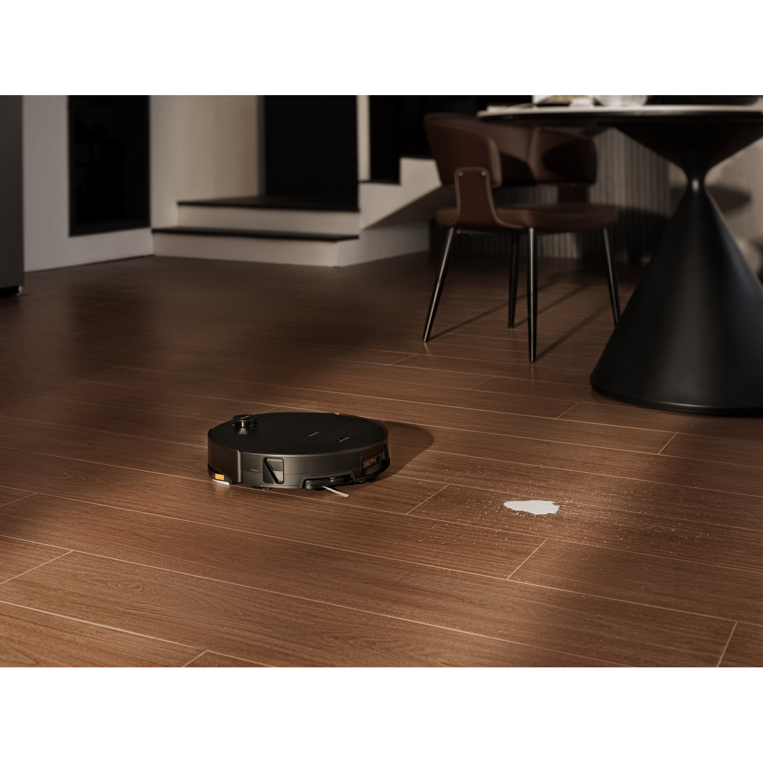 Un robot aspirateur noir nettoie un sol en bois dans une pièce moderne avec table et chaises.