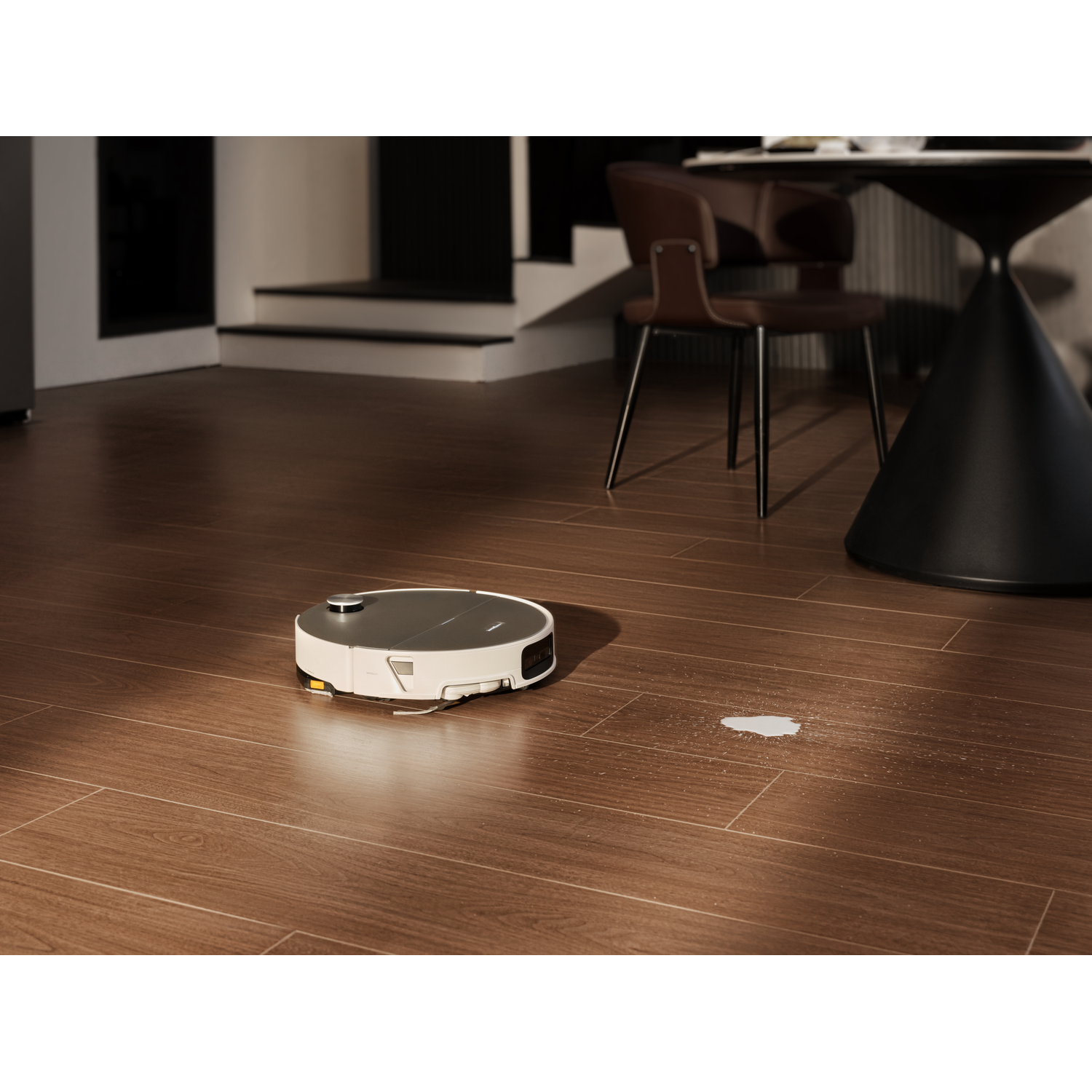 Un robot aspirateur nettoie un sol en bois près d'une tache blanche, avec des chaises et une table en arrière-plan.