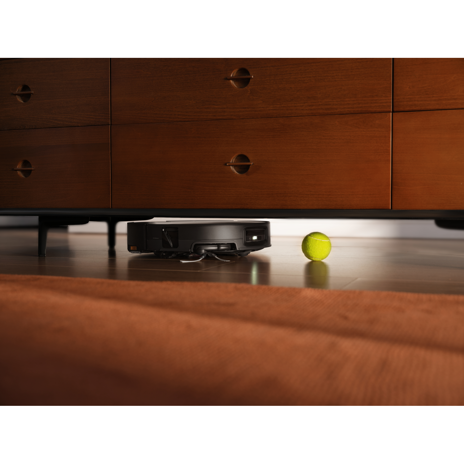 Un robot aspirateur noir nettoie sous une commode en bois près d'une balle de tennis verte.