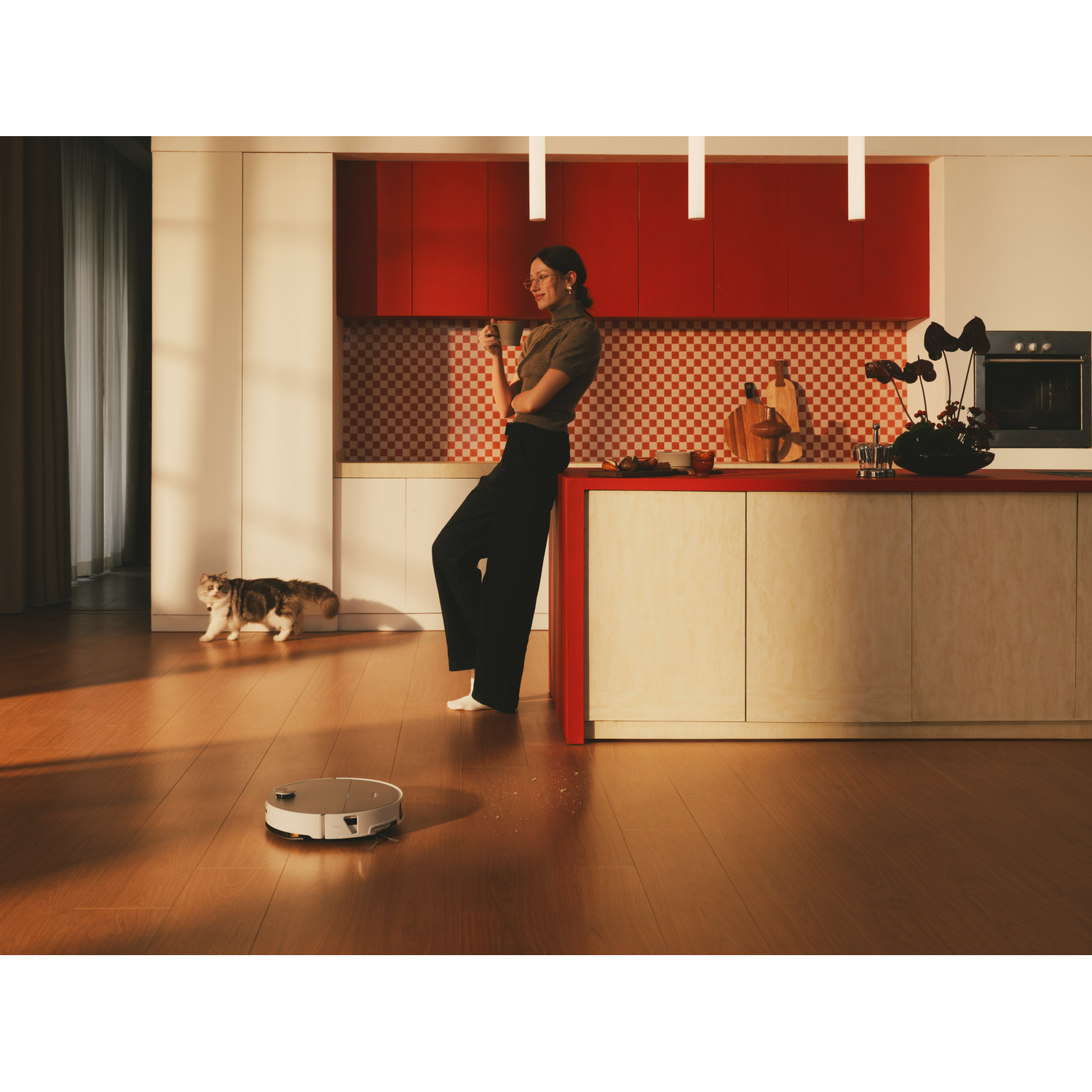 Une femme boit du café dans une cuisine pendant qu'un robot aspirateur nettoie le sol et qu'un chat regarde.