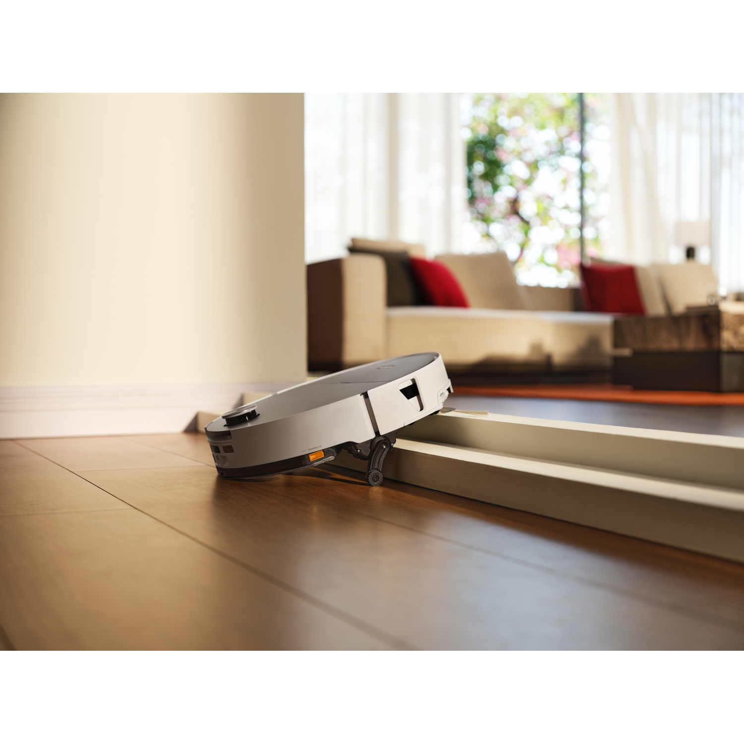 Un robot aspirateur franchit un petit seuil sur un sol en bois dans un salon lumineux.