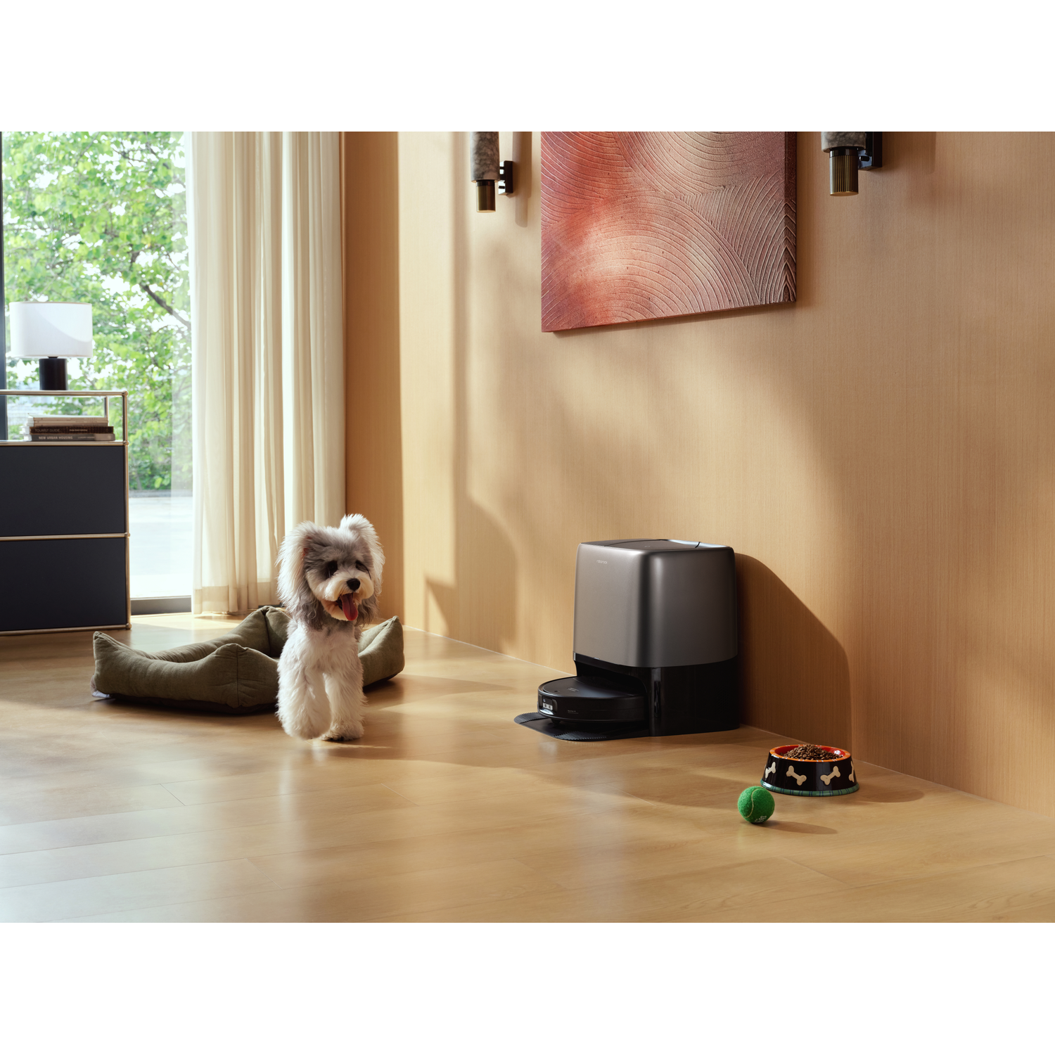 Un petit chien se tient dans le salon près de son lit. Une station d'aspirateur robot est contre le mur.