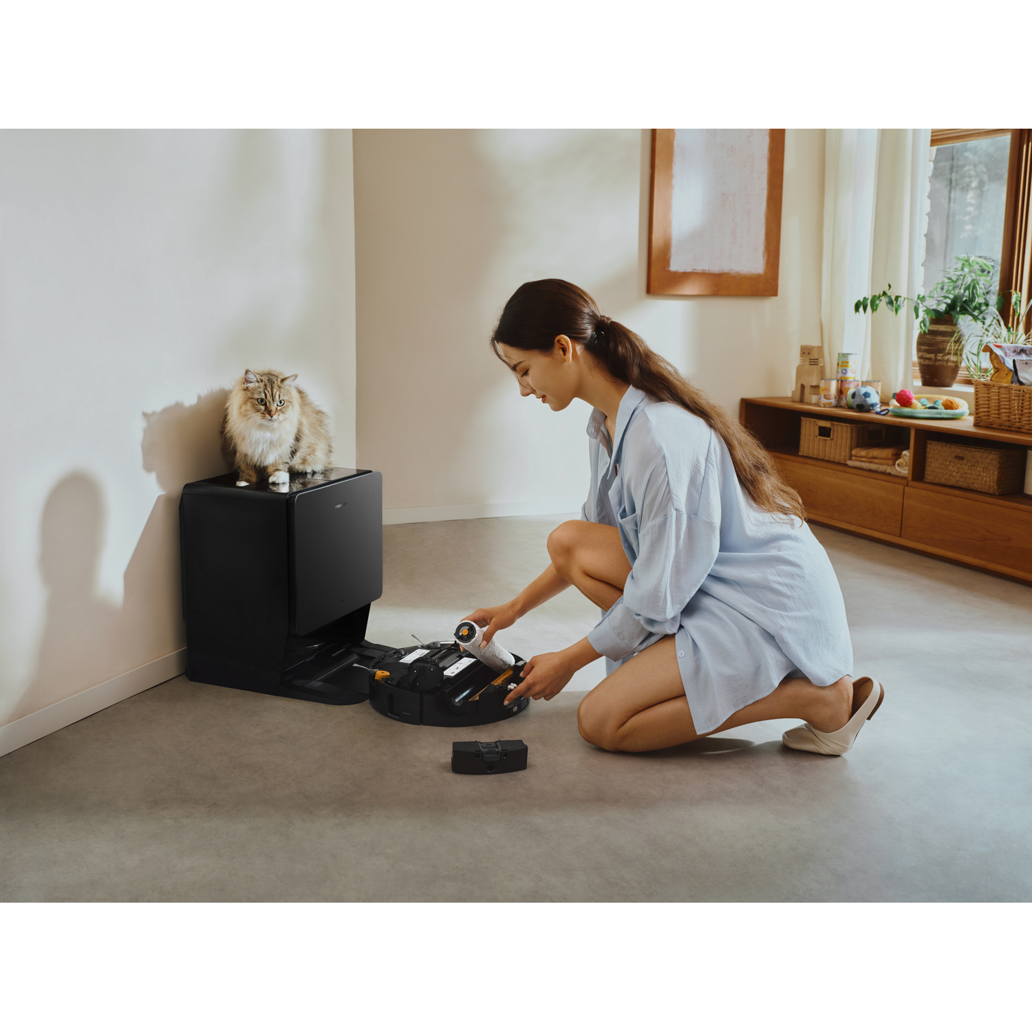 Une femme à genoux entretient un robot aspirateur tandis qu'un chat l'observe depuis une station d'accueil.