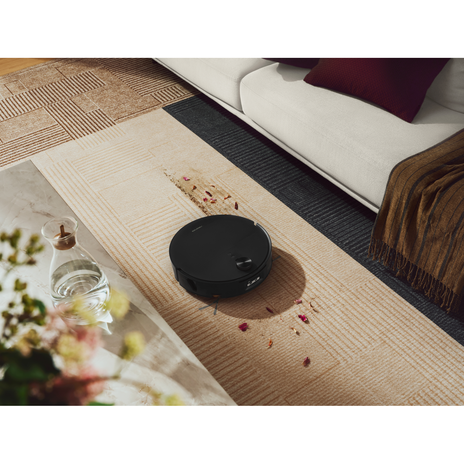 Un robot aspirateur noir nettoie un tapis avec des pétales de fleurs devant un canapé.