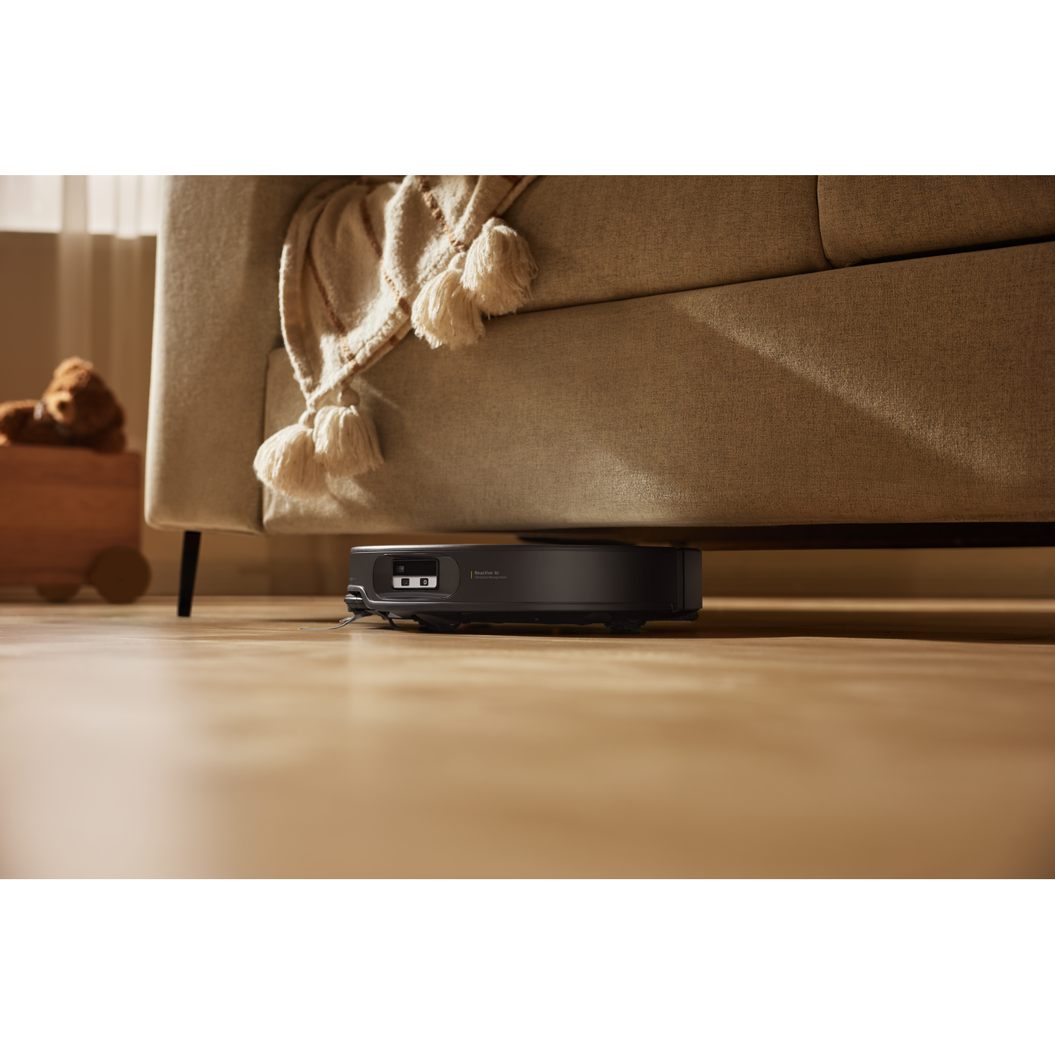 Un robot aspirateur noir nettoie le sol en bois sous un canapé beige avec une couverture.
