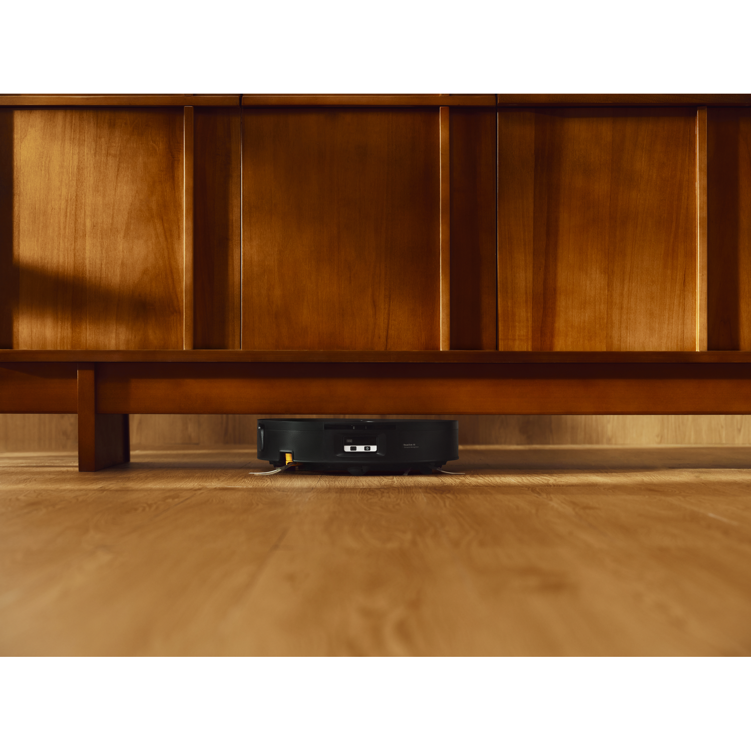 Un robot aspirateur noir nettoie le sol en bois sous un grand meuble en bois marron.