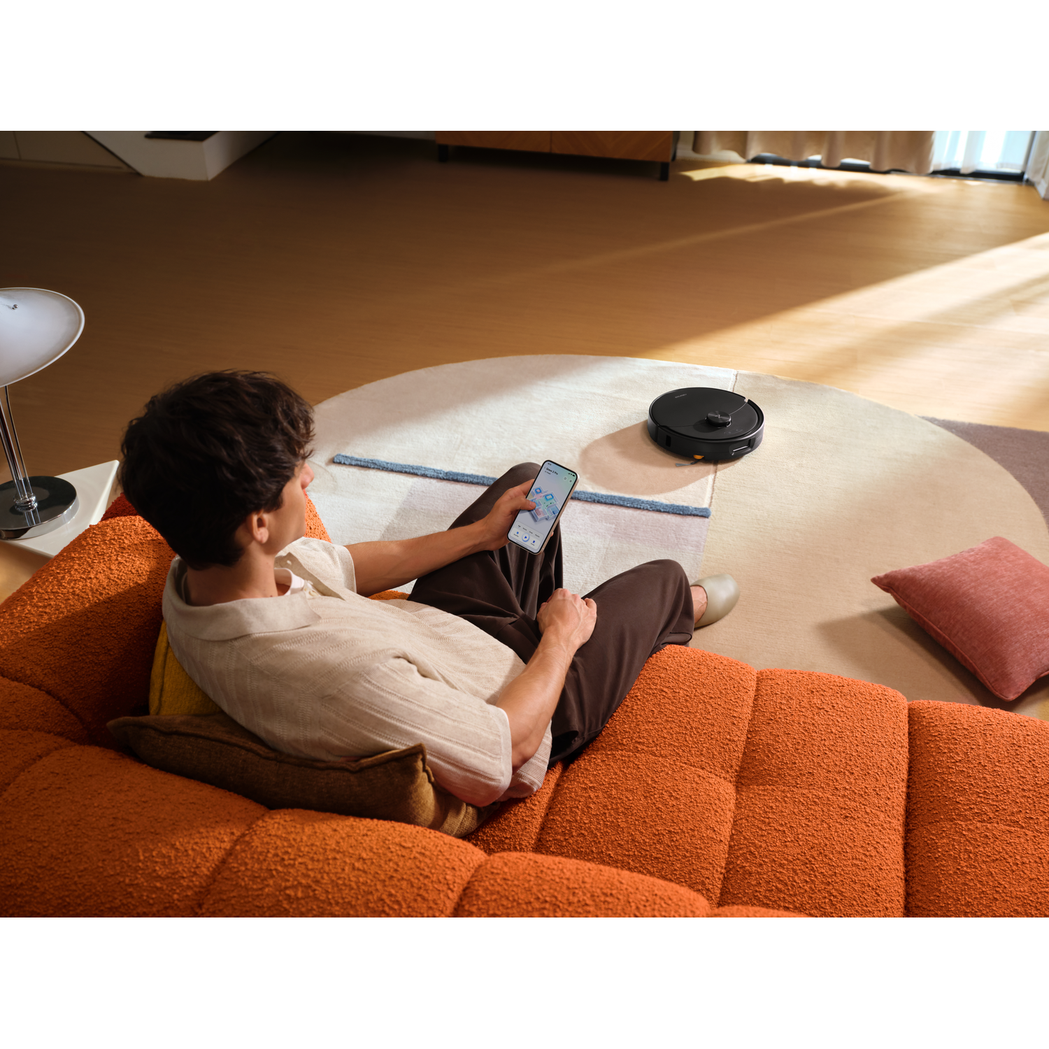Un homme assis sur un canapé orange utilise son smartphone pour contrôler un robot aspirateur.