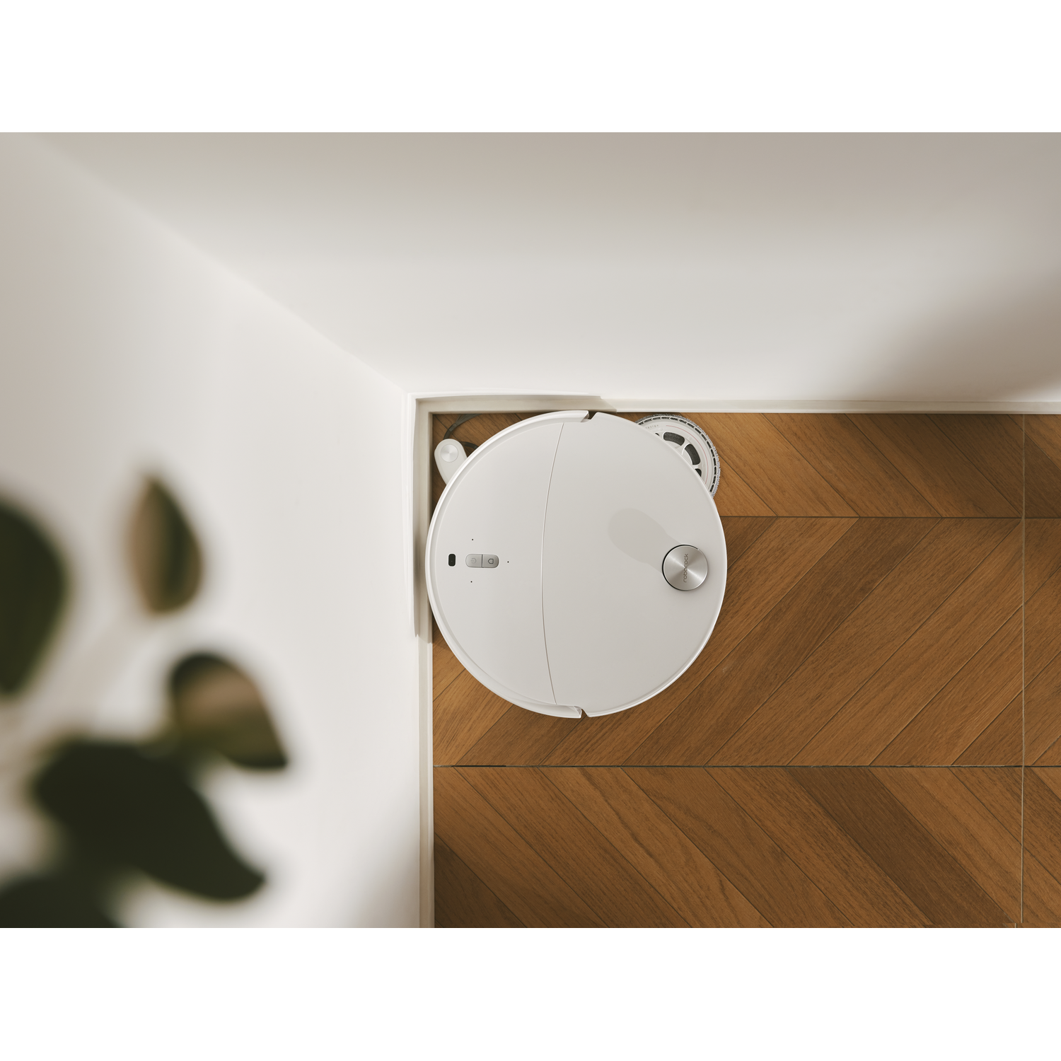 Un robot aspirateur blanc nettoie un coin sur un sol en bois à motifs chevrons.