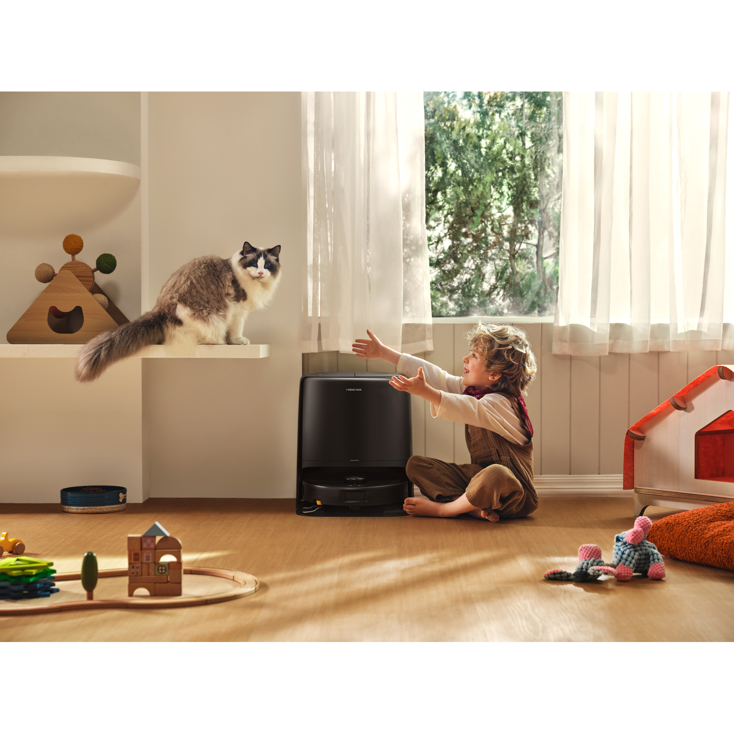 Un garçon assis sur un sol en bois tend la main vers un chat, avec un robot aspirateur noir à proximité.
