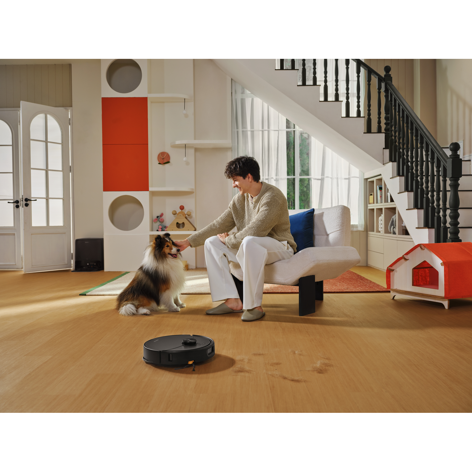 Un homme en pantalon blanc est assis sur une chaise et caresse un chien, tandis qu'un aspirateur robot nettoie le sol.