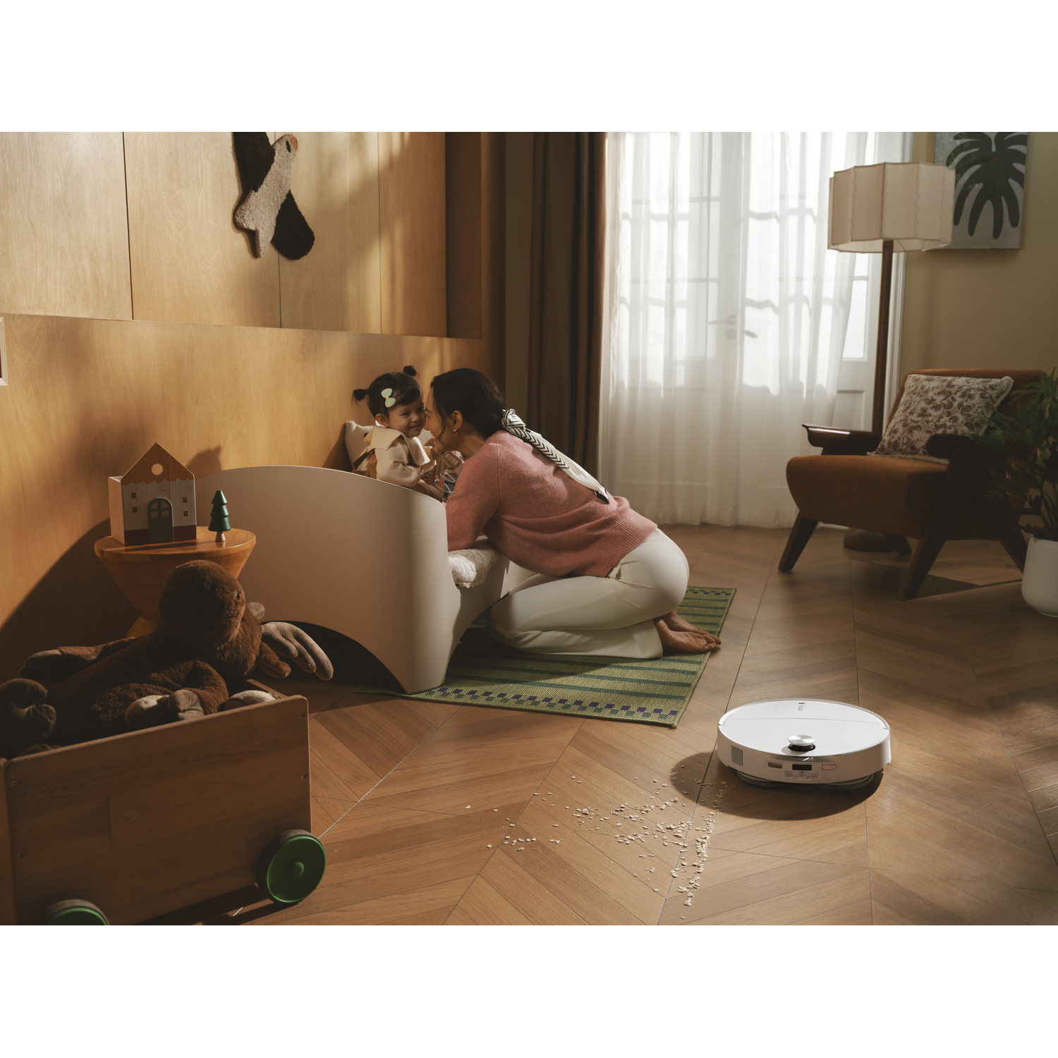 Mère agenouillée près du lit, sourit à l'enfant; robot aspirateur nettoie le sol.