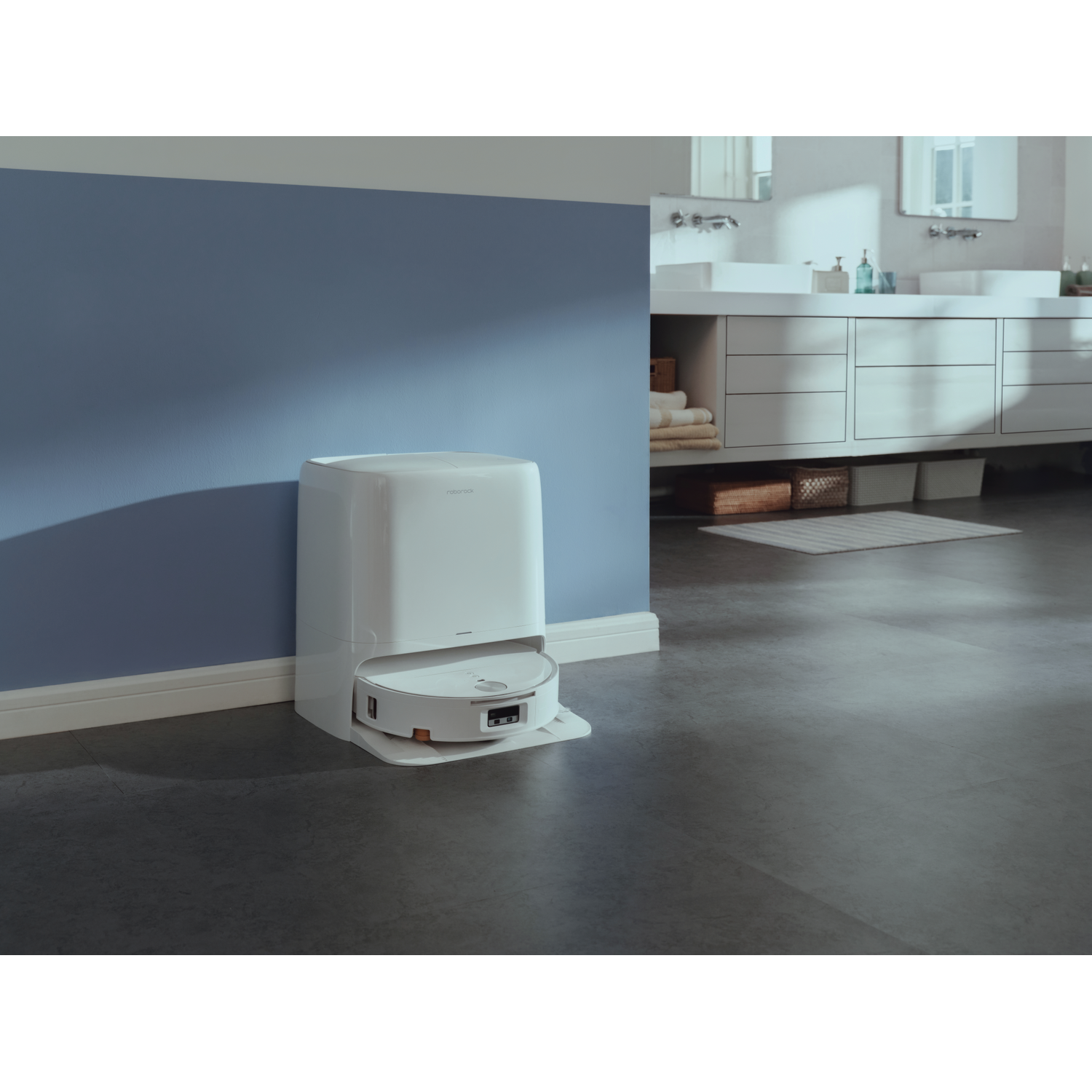 Un robot aspirateur blanc s'amarre à sa station dans une salle de bain moderne avec un mur bleu et des lavabos.