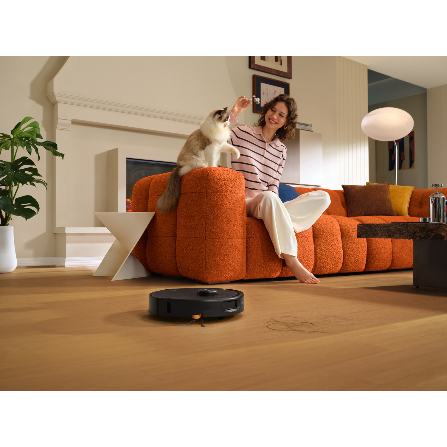 Une femme joue avec son chat sur un canapé orange pendant qu'un robot aspirateur nettoie le sol.