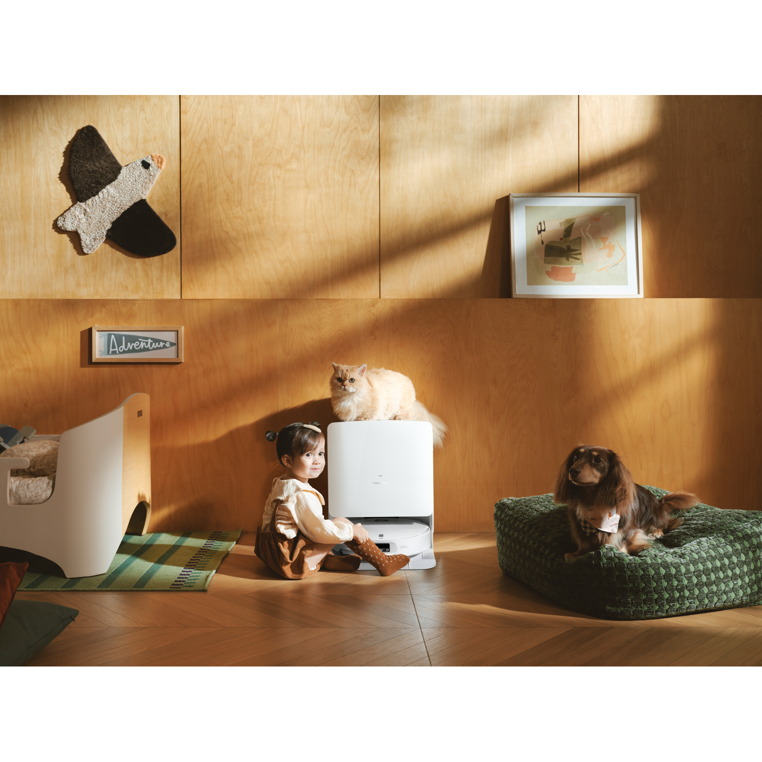Un jeune enfant, un chat et un chien dans une pièce en bois avec une station de robot aspirateur.
