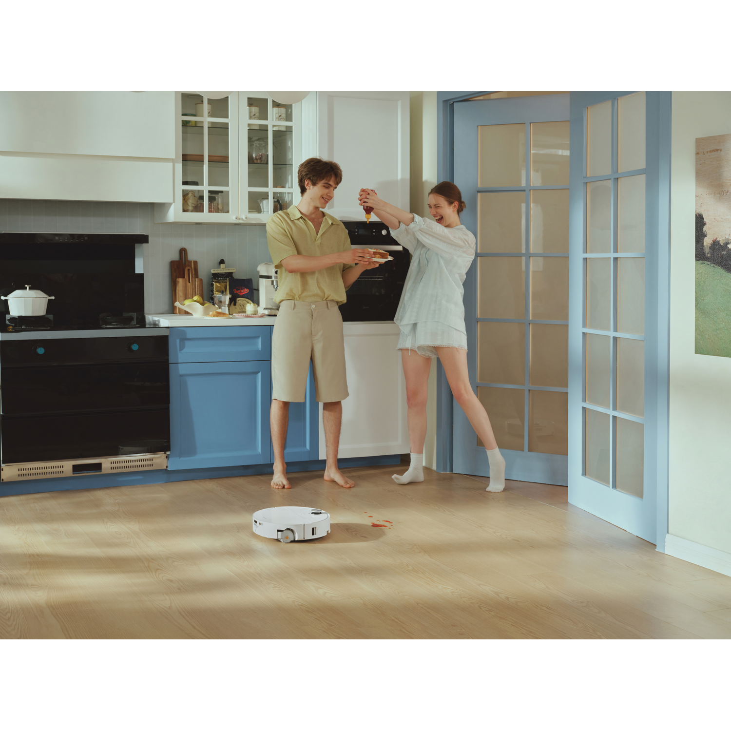 Un couple souriant dans une cuisine joue avec de la nourriture pendant qu'un robot aspirateur nettoie une tache.