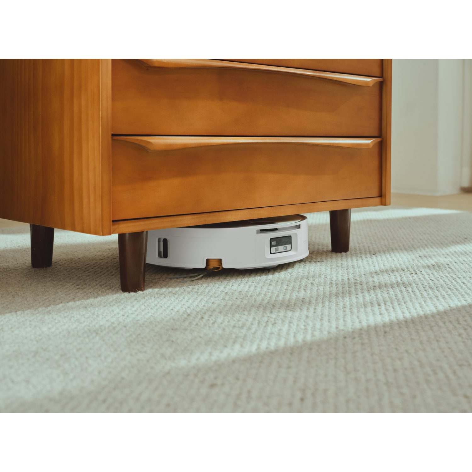 Un robot aspirateur blanc nettoie sous une commode en bois sur un tapis clair.