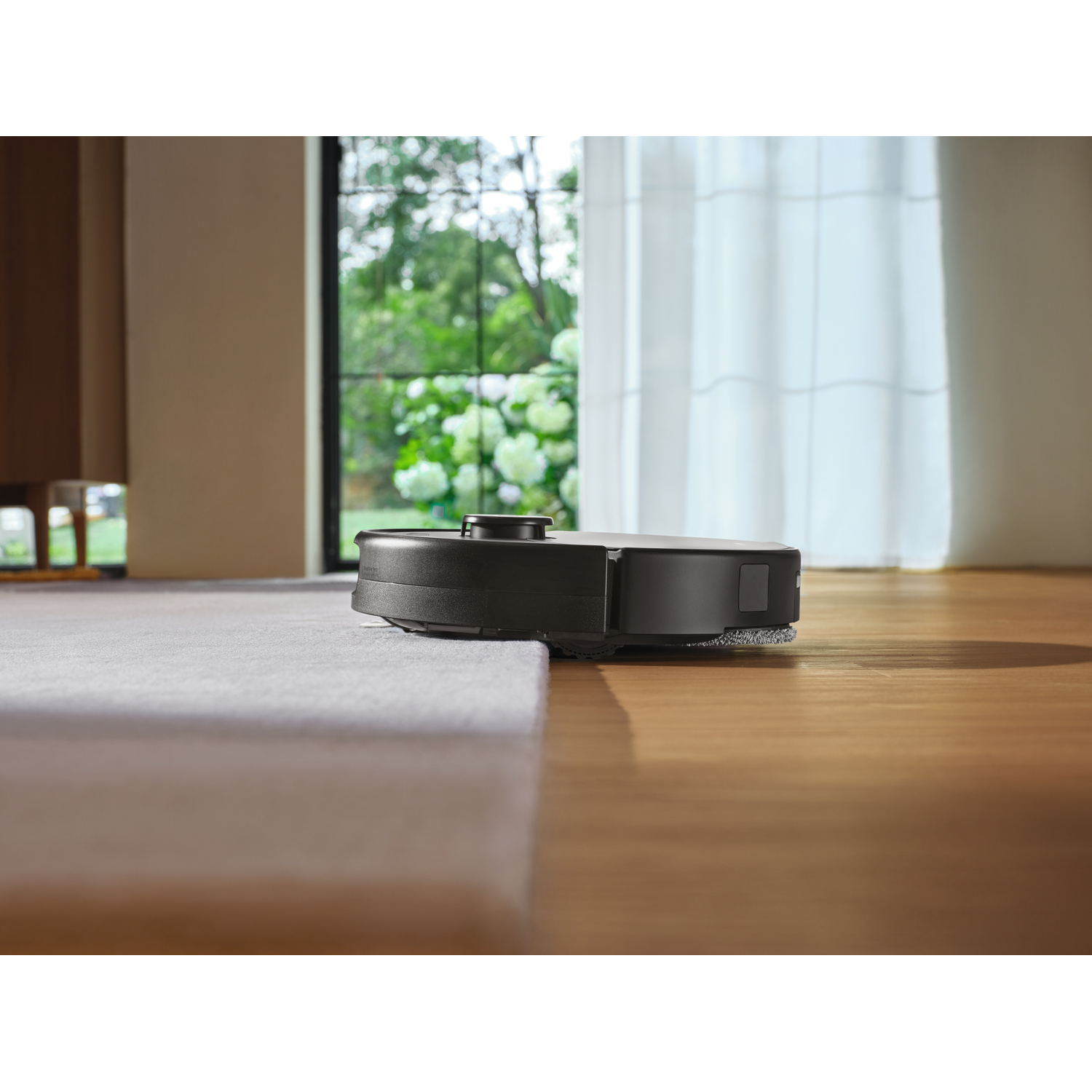 Un aspirateur robot noir nettoie la transition entre un tapis et un plancher en bois dans une pièce ensoleillée.