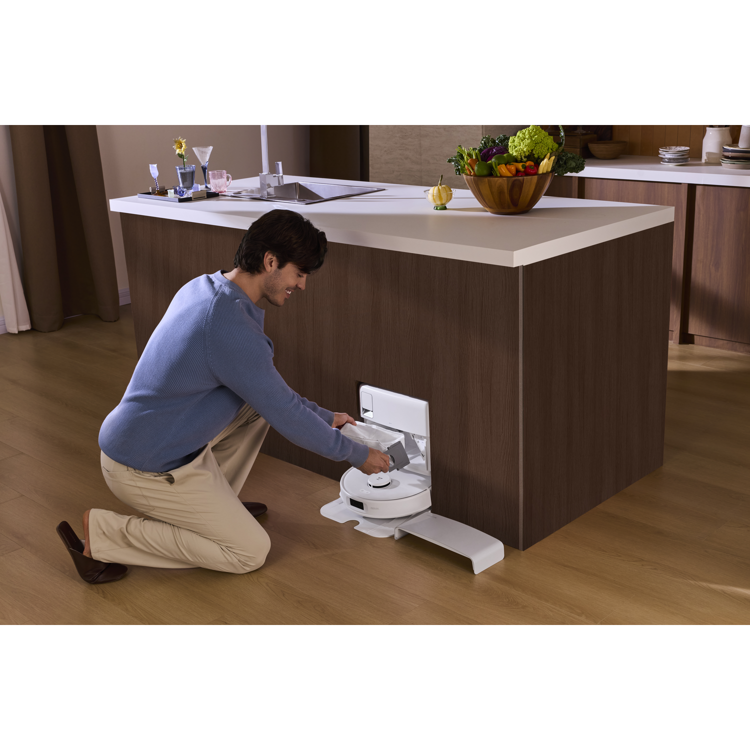 Un homme genouillé près d'un îlot de cuisine interagit avec un robot aspirateur blanc.