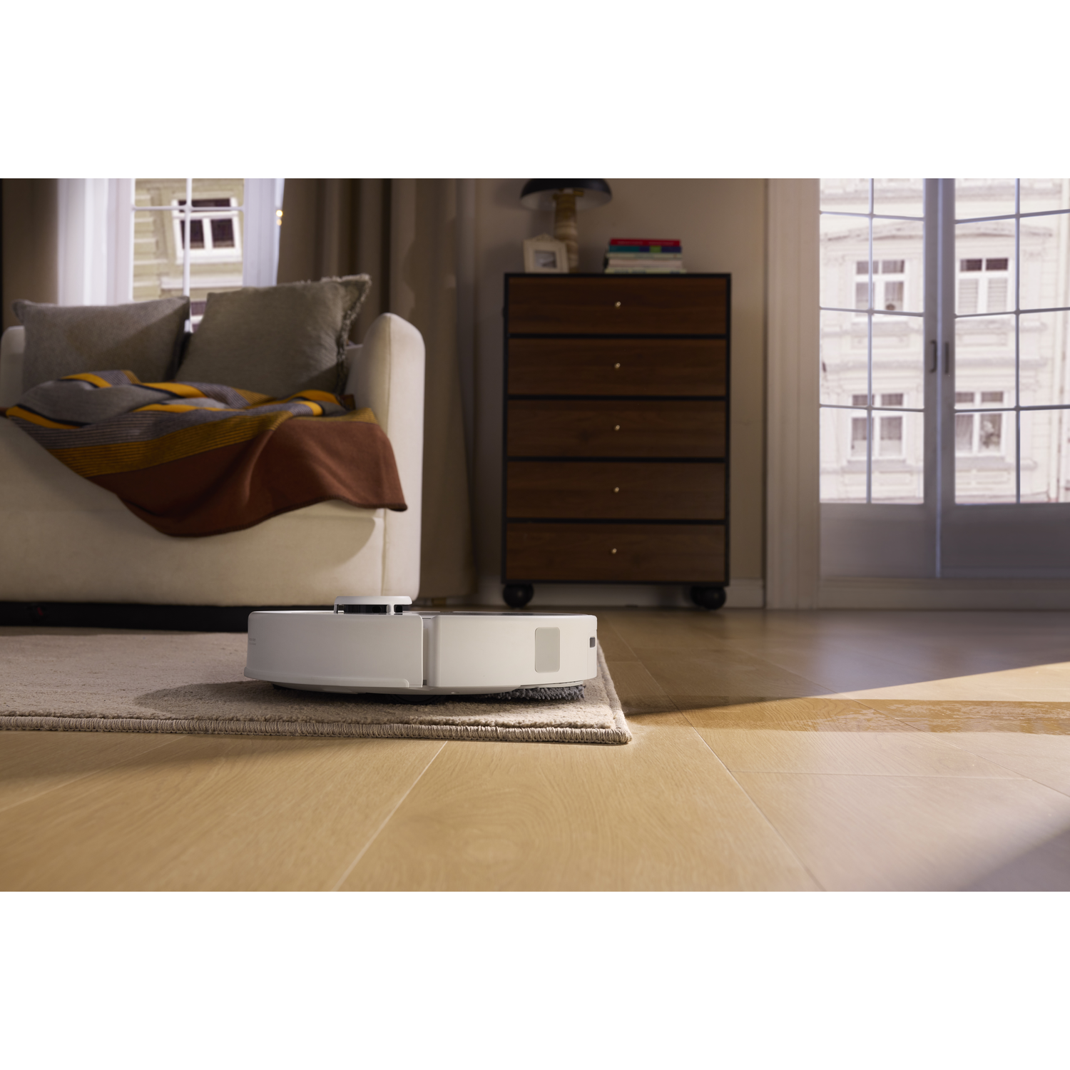 Un robot aspirateur blanc nettoie le sol d'un salon près d'un tapis, d'un canapé et d'une commode.