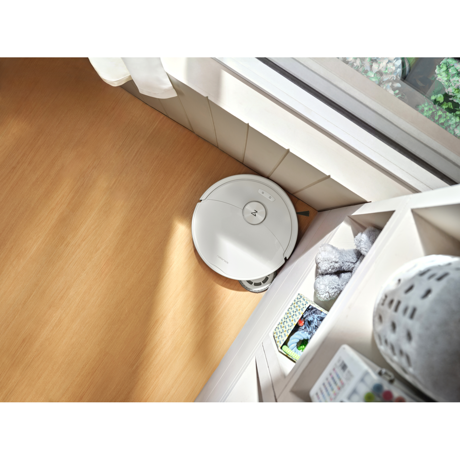 Un robot aspirateur blanc nettoie un plancher en bois à côté d'un meuble et d'une fenêtre.