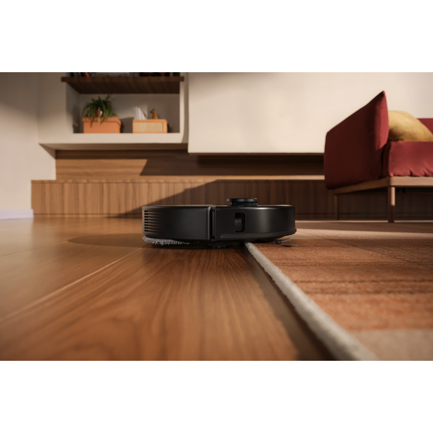 Un aspirateur robot noir nettoie un sol en bois à côté d'un tapis dans un salon moderne.