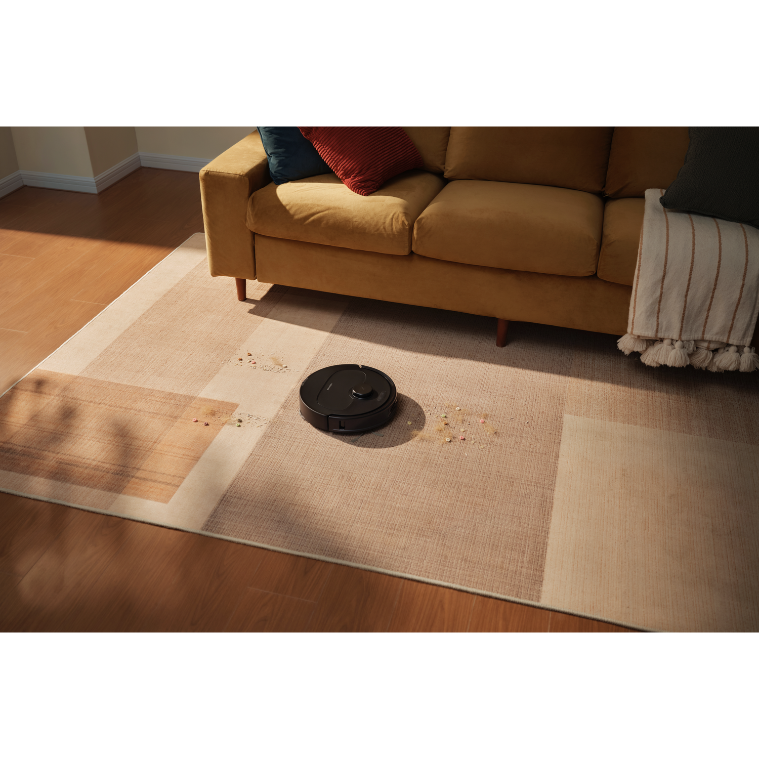Un aspirateur robot noir repose sur un tapis géométrique devant un canapé jaune, près de débris.