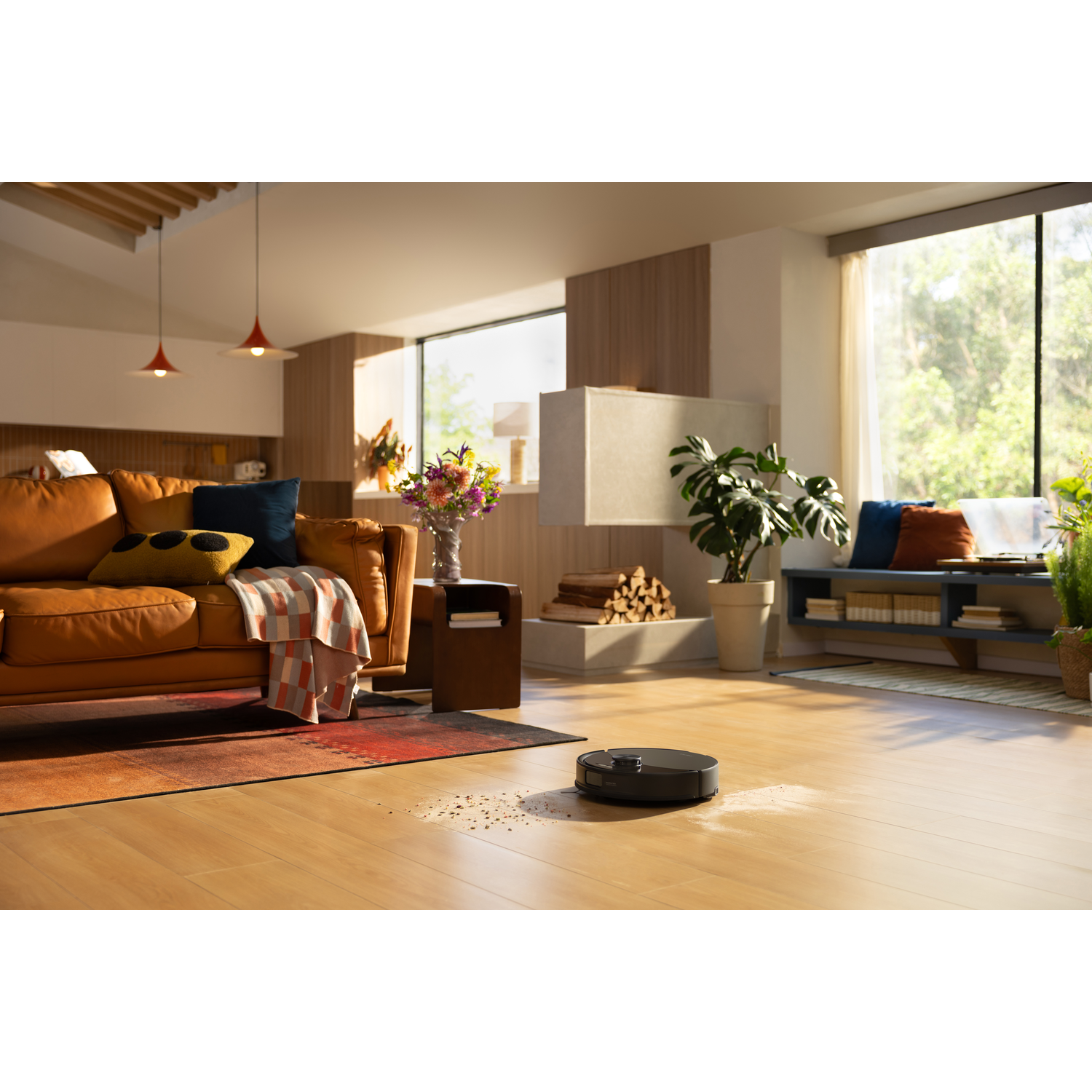 Un robot aspirateur nettoie un sol en bois dans un salon moderne et ensoleillé avec canapé en cuir et cheminée.