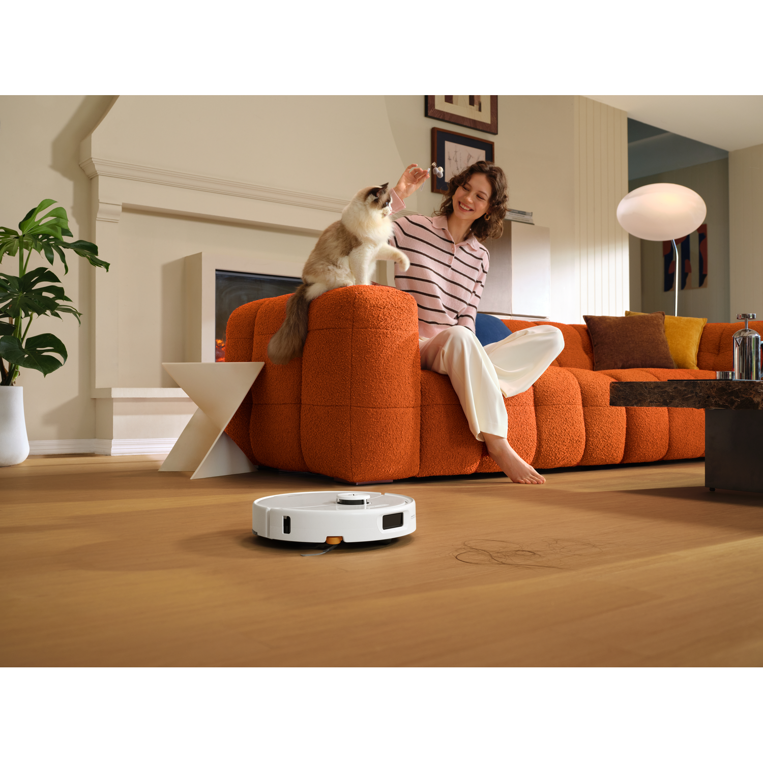 Un robot aspirateur nettoie le sol en bois près d'une femme jouant avec son chat sur un canapé orange.